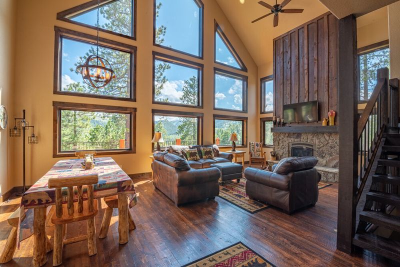 Spacious mountain lodge-style living and dining area with vaulted ceilings, floor-to-ceiling windows offering forest views, leather furniture, stone fireplace, and rustic wood accents; custom home by Legacy Custom Homes & Design in Spearfish, SD.