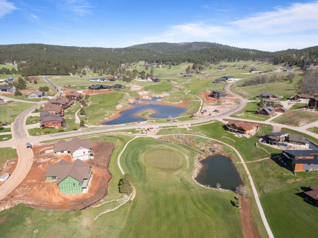 Drone view of Apple Springs housing development near Sturgis, SD, showcasing homes under construction, curving roads, ponds, and a golf course surrounded by forested hills.
