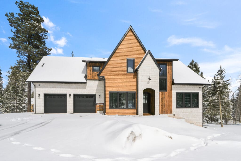 Exterior of The Cottage custom home in Powder House Pass Subdivision, Lead, featuring a snowy landscape. The home blends rustic wood accents with light-colored stone, large windows, and modern architectural details. The scene is set in a serene winter environment with snow-covered ground and trees.