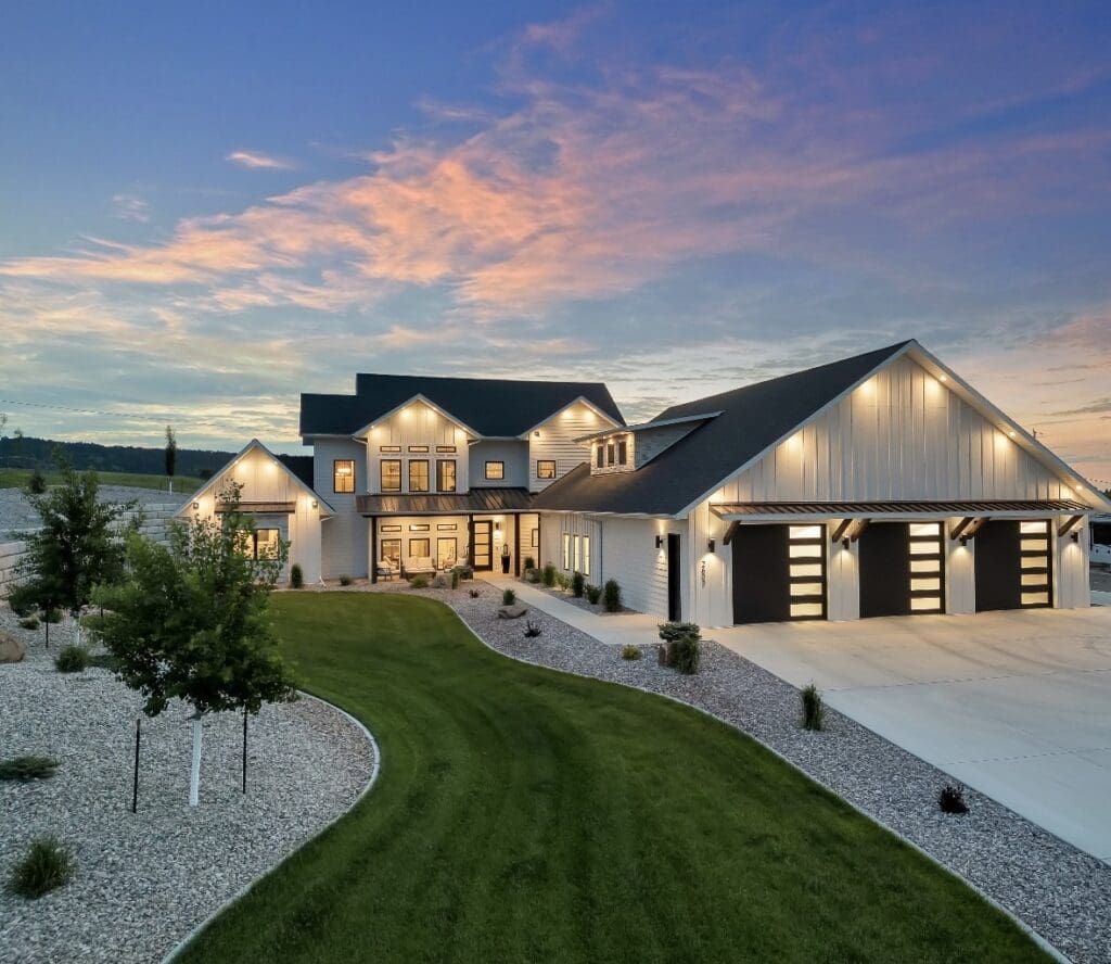 Modern custom home in Top Shelf Development built by Legacy Custom Homes & Design in Spearfish, featuring large windows, a three-car garage, professional landscaping, and exterior lighting at sunset.