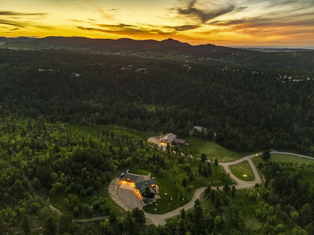 Aerial sunset view of a luxury custom home built by Legacy Custom Homes & Design in Pillar Peak Estates, Deadwood, South Dakota. The mountain residence sits on a large wooded lot surrounded by Black Hills forest, rolling terrain, private roads, and panoramic views of the Deadwood landscape at dusk.