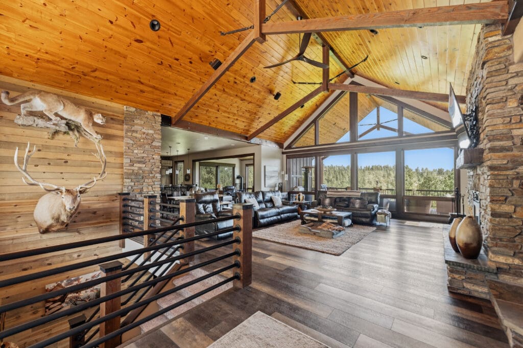 Luxury custom home interior built by Legacy Custom Homes & Design in Deadwood, SD, featuring vaulted tongue-and-groove wood ceilings, exposed timber beams, floor-to-ceiling windows, stone fireplace, and panoramic Black Hills views.