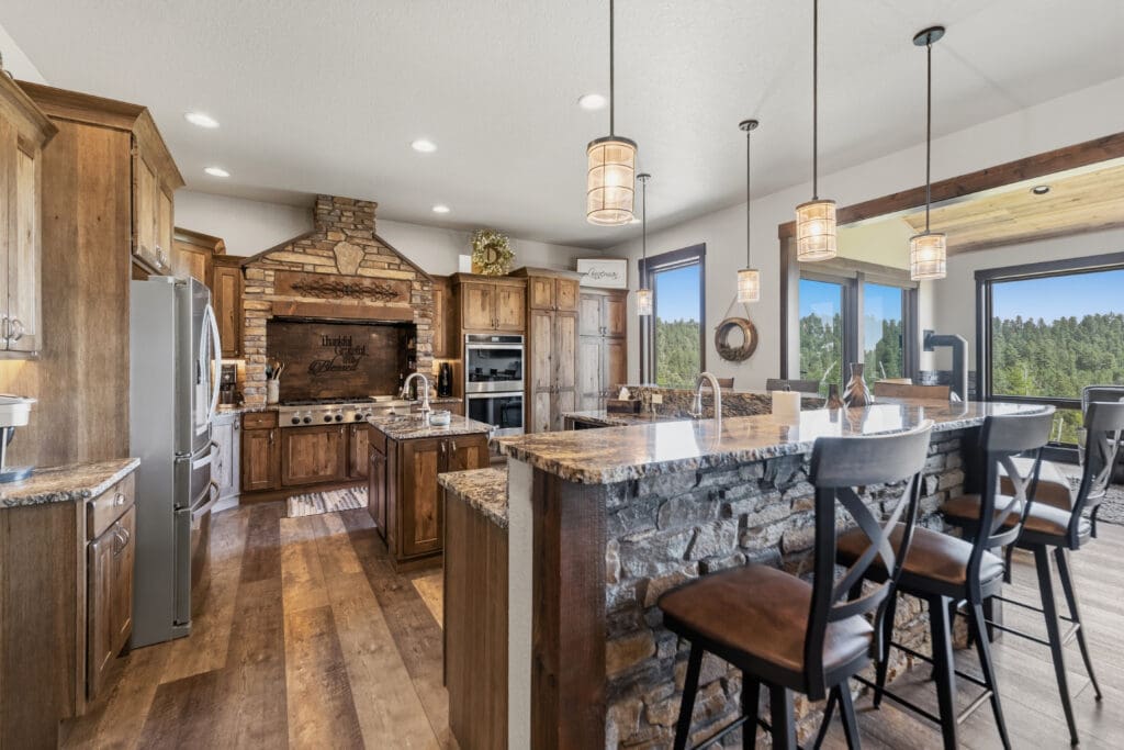 Luxury custom kitchen built by Legacy Custom Homes & Design in Deadwood, SD, featuring custom wood cabinetry, natural stone accents, granite countertops, oversized island seating, and expansive Black Hills views.